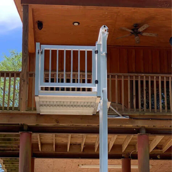 Outdoor home elevator on a wooden house, connecting upper balcony to the ground. White metal framework and wooden ceiling fan visible.