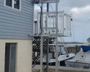 Outdoor home elevator next to a blue house by a marina, boats docked nearby under a cloudy sky.