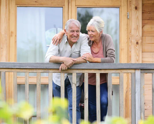 Elderly couple smiling on wooden deck Elderly couple smiling on wooden porch, leaning on railing, with a cozy wooden house in the background.