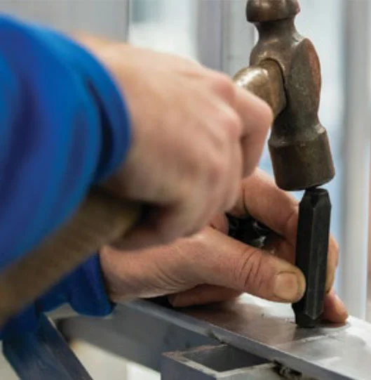 Hands hammering a chisel into a metal bar, possibly part of a home elevators installation process.